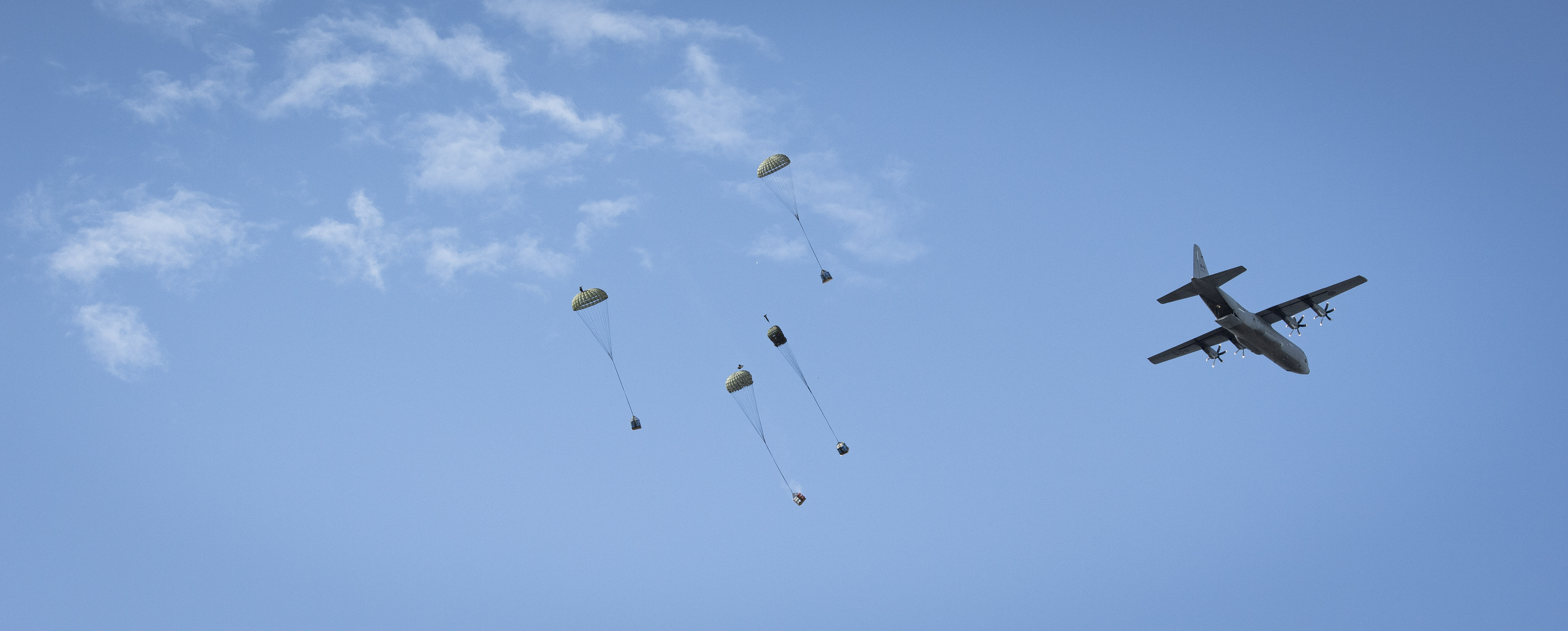Four containers attached to parachutes falling in the sky with a large grey propeller aircraft flying overhead on the right.