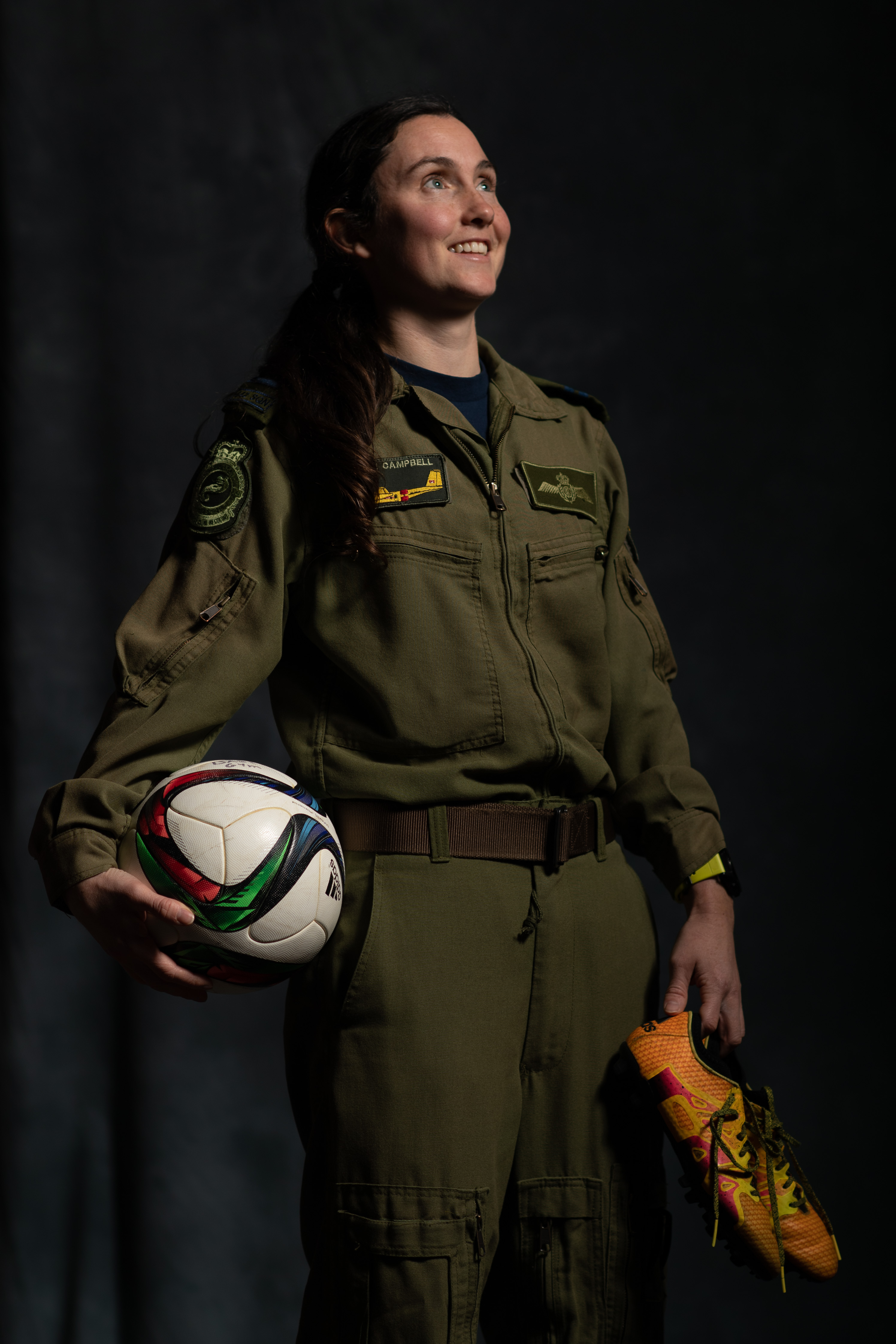 A smiling woman wearing an olive-green flight suit and holding a ball and sports shoes.