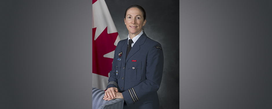 diapositives - Une femme souriante vêtue d’un uniforme militaire bleu se tient devant un drapeau du Canada, ses mains reposant sur une chaise bleue.